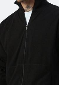 Giacca nera con zip, colletto alto, texture morbida e cuciture piatte. Presenta un tirante in metallo argentato e un design a strati.