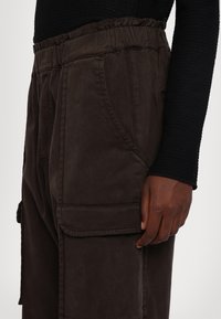 Pantalon cargo marron foncé avec grande poche latérale et taille élastique, porté avec un haut noir à manches longues, main reposant sur le côté.