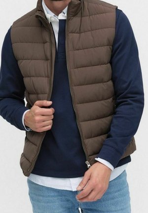 Homme portant un gilet matelassé marron par-dessus une chemise bleu marine à manches longues et un jean bleu clair, tenant la fermeture éclair du gilet d'une main.