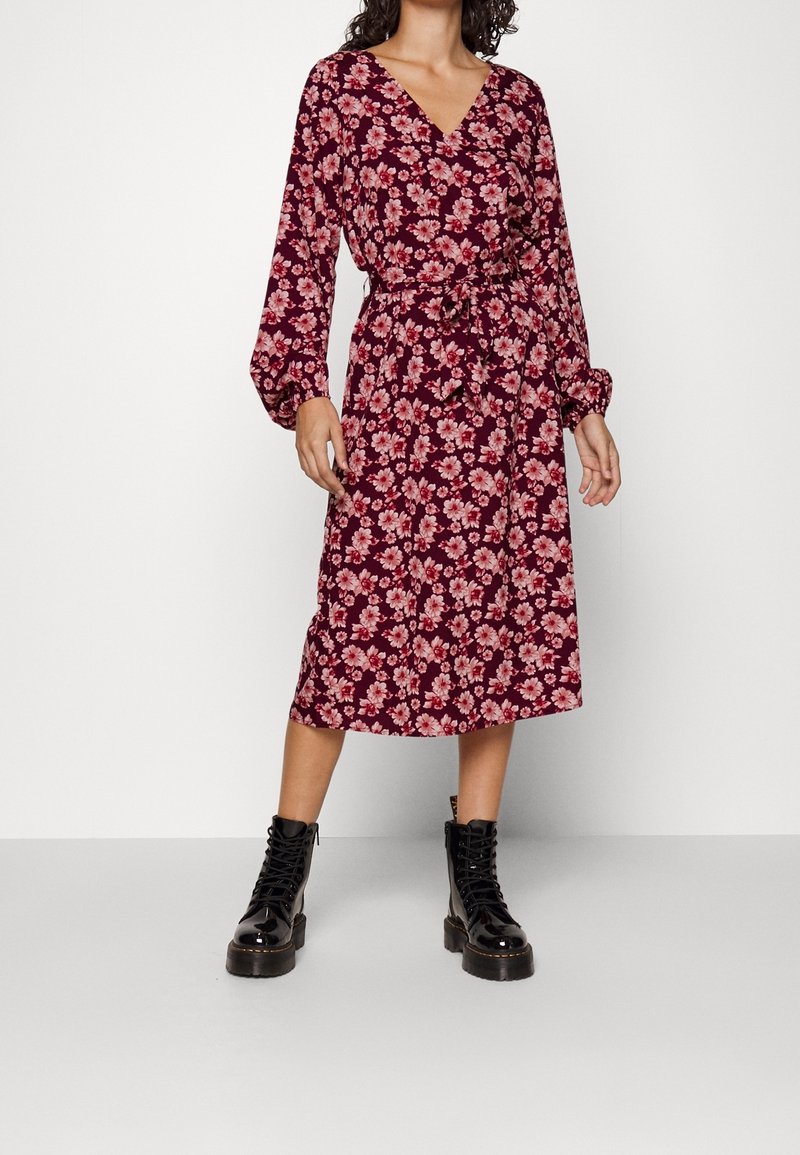 Femme portant une robe midi florale bordeaux à manches longues et taille nouée, associée à des bottines noires brillantes à lacets.