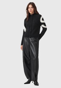 Maglione zip-up nero con motivo bianco sulle maniche, realizzato in materiali morbidi. Abbinato a pantaloni in pelle nera larghi e scarpe nere a punta.