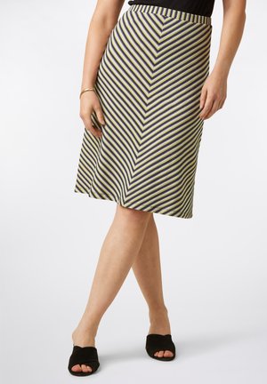 Femme portant une jupe mi-longue à motifs rayés diagonaux et des talons ouverts noirs, debout avec les jambes croisées sur un fond blanc.