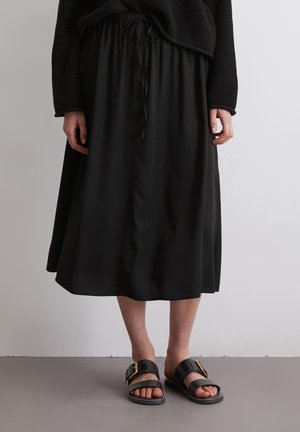 Osoba nosząca czarną spódnicę midi ze ściągaczem w talii oraz czarne sandały ze złotymi klamrami, stojąca na szarej podłodze przy białej ścianie.
