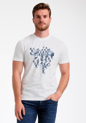 T-shirt bianco a girocollo realizzato in morbido cotone, con un design grafico geometrico blu e grigio sulla parte anteriore, indossato con pantaloni scuri.
