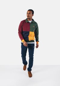 Värgblöckt fleece-pullover i bourgogne, grön, marinblå och gul; kvartdragkedja; ihopparad med blå byxor och bruna mockaskor.