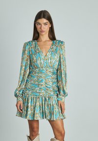 Mujer con un vestido de manga larga, escote en V, de patrón turquesa y marrón con cintura fruncida y falda con volantes, de pie frente a un fondo liso.