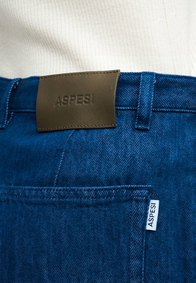 Κοντινό πλάνο μπλε τζιν με καφέ δερμάτινη ετικέτα και λευκή υφασμάτινη ετικέτα, και οι δύο με ανάγλυφο το όνομα της μάρκας «ASPESI».