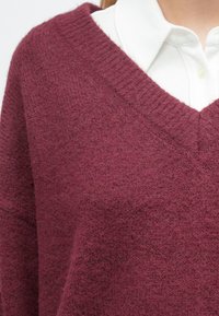Bordový sveter s výstrihom do v tvaru V, vyrobený z mäkkej, štruktúrovanej tkaniny. Má rebrované okraje na výstrihu a manžetách, vrstvený cez bielu košeľu s golierom.