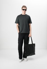 Black leather tote bag held by a person wearing a dark gray t-shirt and black cargo pants, complemented by white sneakers and sunglasses.