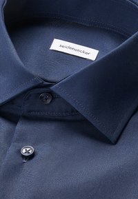 Marineblaues Hemd mit spitzem Kragen und zwei dunklen Knöpfen, mit einem weißen Etikett mit dem Markennamen "seidensticker" im Krageninneren.