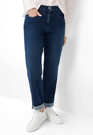 Person trägt dunkelblaue hochgekrempelte Jeans, weiße Turnschuhe und ein hellrosa Langarmshirt, eine Hand in der Tasche.