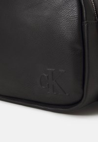 Borsa nera in pelle con una superficie testurizzata, dotata di un logo "CK" in rilievo. Ha una forma rettangolare elegante e tocchi di design discreti.