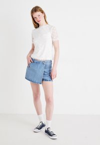 Hvit blonde topp med korte ermer og bølgete kanter, kombinert med en blå denim omslagskjørt som har en frontlomme og fransede kanter.