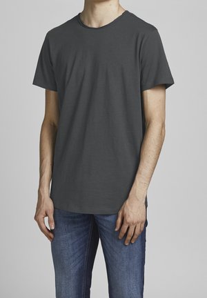 Homme portant un t-shirt uni gris foncé à manches courtes et un jean bleu, debout devant un fond gris clair.