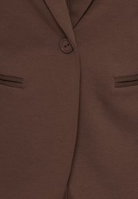 Gros plan de l'avant d'un blazer marron avec un seul bouton marron fermé et deux poches passepoilées horizontales étroites.