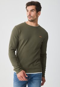 Άνδρας με ελιά πράσινο μπλουζάκι μακρυμάνικο με μια μικρή καφέ λογότυπη ετικέτα και μπλε τζιν, κοιτάζοντας προς τα αριστερά σε απλή φόντο.