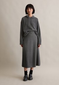 Grau gestrickter Kapuzenpullover mit einer Vorderstasche und Kordelzug, kombiniert mit einem passenden langen Rock. Das Model trägt schwarze Plateauschuhe und gerippte Socken.