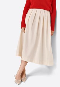 Jupe midi plissée crème en tissu lisse, associée à des chaussures à talons beiges à bout pointu, avec un pull rouge à côtes sur le dessus.