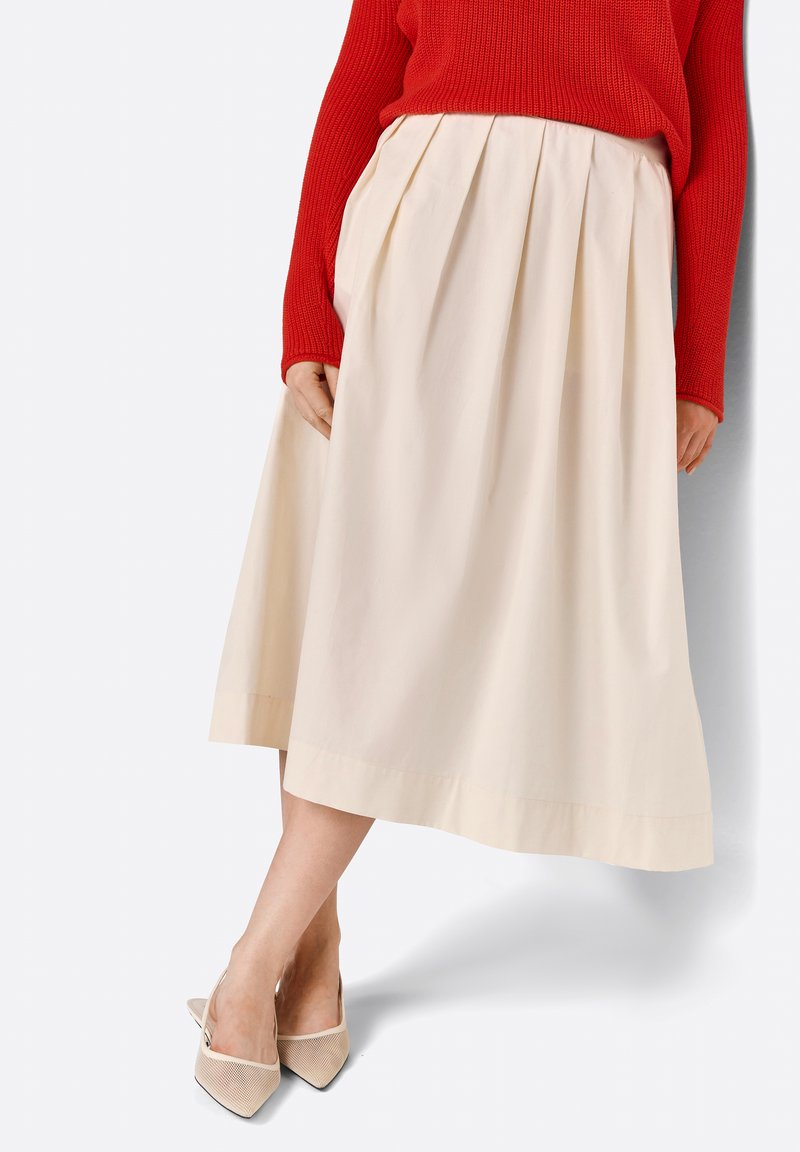 Jupe midi plissée crème en tissu lisse, associée à des chaussures à talons beiges à bout pointu, avec un pull rouge à côtes sur le dessus.
