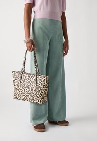 Sac cabas imprimé léopard avec deux anses, forme structurée, motif beige et noir. Porté avec un pantalon large en velours côtelé vert clair et des chaussures marron.