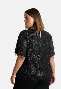 Svart paljettopp med fransdesign, korta ärmar och en nyckelhålsöppning bak. Texturerad med glänsande, vertikala paljettaccenter.