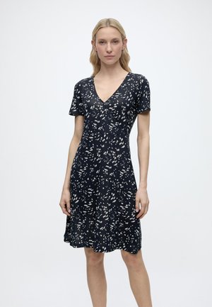 Nő fekete, rövid ujjú ruhában, amelyen fehér absztrakt foltok vannak, V-nyakú és térdig érő, egyszínű fehér háttér előtt áll.