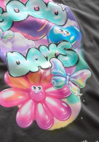 Grafisk t-shirt med en kolagigrå bakgrund som har färgglad, glansig text som säger "Bra Dagar" samt tecknade blommor och fjärilar.