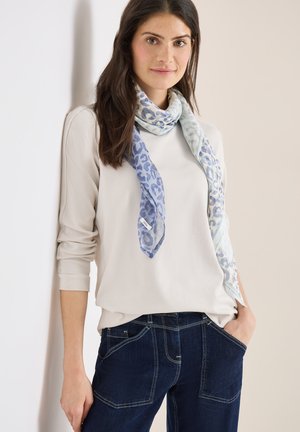 Femme portant un haut clair à manches longues, une écharpe bleue à motifs, et un jean bleu foncé, debout avec une main dans la poche, sur un fond clair.