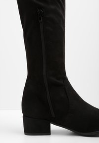Schwarze Wildleder-Stiefel bis zum Knie mit niedrigem Blockabsatz, seitlichem Reißverschluss und glatter Textur, die subtile Sch stitching-Details entlang der Nähte aufweisen.