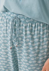 Svetle modré loungewear šortky s bílým vzorem mušlí, vybavené elastickým pasem a řetízkem se červenými akcenty. Měkká, prodyšná tkanina.