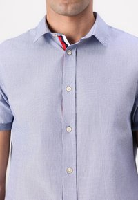 Camisa de manga corta azul claro con botones, confeccionada en un tejido texturizado, con cuello interior a rayas en contraste y botones crema.