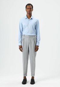 Chemise bleu clair à manches longues, boutonnée, associée à un pantalon gris taille haute avec un design plissé et deux boutons latéraux. Chaussures noires.