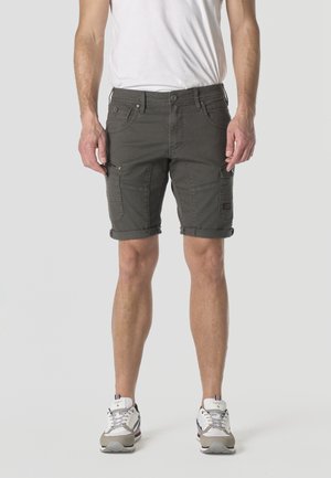 Hombre con pantalones cortos cargo gris oscuro con dobladillos enrollados, camiseta blanca y zapatillas blancas, de pie contra un fondo claro y liso.