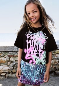 Schwarzes, kurzes T-Shirt mit pink-türkisfarbigem Grafikdesign, kombiniert mit einem Paillettenrock in Farbverlauf von Blau bis Lila.