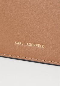 Lys brun lærflate med en teksturert finish. Gullpräget "KARL LAGERFELD"-logo og kontrasterende sømdetaljer langs kanten.