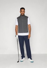 Γκρι μπουφάν χωρίς μανίκια με ψηλό γιακά, λευκή κοντομάνικη μπλούζα, ναυτικές παντελόνες και λευκά παπούτσια γκολφ. Κρατάει μια βέργα γκολφ με ένα γάντι σε ένα χέρι.