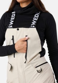 Beige vandtætte overaller med justerbare sorte stropper, der har "SWEDEN" tekst, lynlåslommer og en struktureret sort langærmet bluse under.