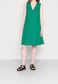 Zielona sukienka bez rękawów z dekoltem w szpic. Wykonana z lekkiego materiału, o kroju w literę A. Zestawiona z czarnymi sandałami na paski.