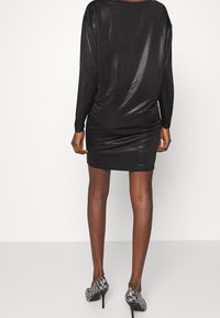 Svart, blänkande långärmad klänning med figurnära silhuett och samlad midjedetalj. Matchat med mönstrade svarta klackar.