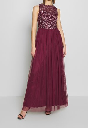 Bordeauxrotes Abendkleid mit einem perlenverzierten Oberteil und ärmellosem Design. Fließender Tüllrock reicht bis zum Boden. Schlichte schwarze Sandaletten mit Absatz.