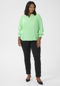 Blouse vert clair avec un décolleté en V, des manches bouffantes et des poignets élastiques. Associée à un pantalon noir ajusté et des talons pointus noirs.