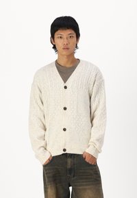 Cardigan en maille crème clair avec un col en V, présentant un motif texturé, des poignets côtelés et cinq boutons foncés à l'avant.