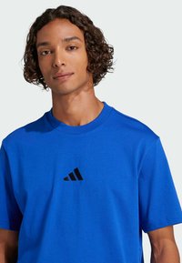 Giovane con capelli ricci che indossa una maglietta Adidas blu brillante con un logo nero sul petto, rivolto in avanti con un'espressione neutra.