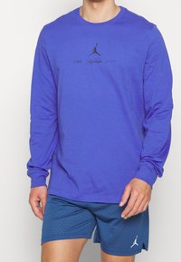 Långärmad lila skjorta i bomull, med en svart grafisk logotyp i mitten. Pairs med blå perforerade shorts.