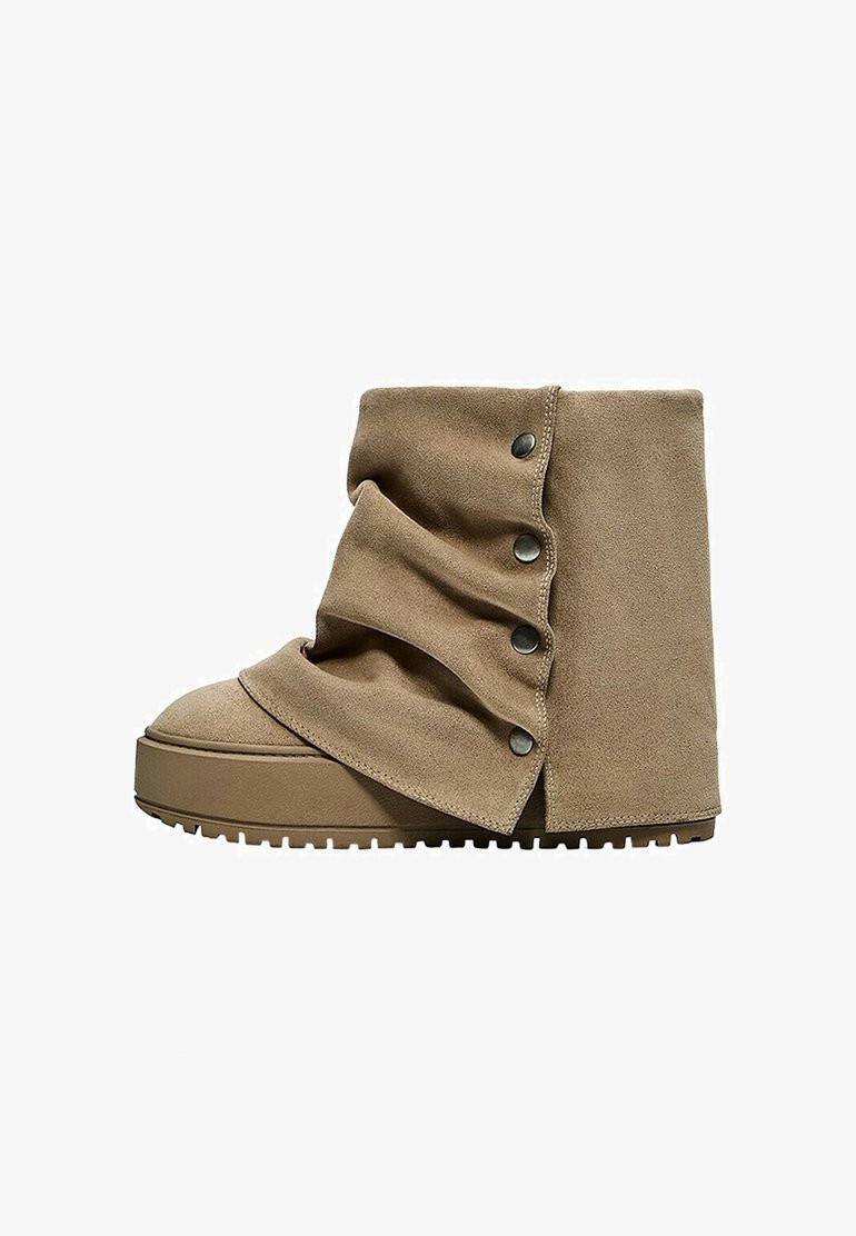 Bottine beige en daim avec un design plié et des boutons-pression métalliques sur le côté, avec une semelle épaisse en caoutchouc avec des crampons.