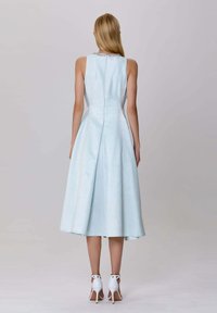 Une robe sans manches bleu clair avec un corsage ajusté et une jupe évasée. Elle présente un tissu texturé et un décolleté décoratif. Des talons blancs à bride cheville.