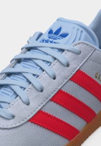 Ανοιχτό γκρι αθλητικό παπούτσι Adidas Gazelle με μπλε κορδόνια, μπλε λογότυπο στη γλώσσα, κόκκινες ρίγες στο πλάι και σόλα χρώματος καραμέλας.