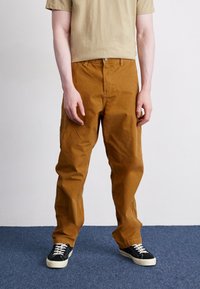 Pantaloni in twill di cotone marrone con gamba dritta, dotati di chiusura a bottone e tasche laterali, abbinati a sneakers nere e bianche.