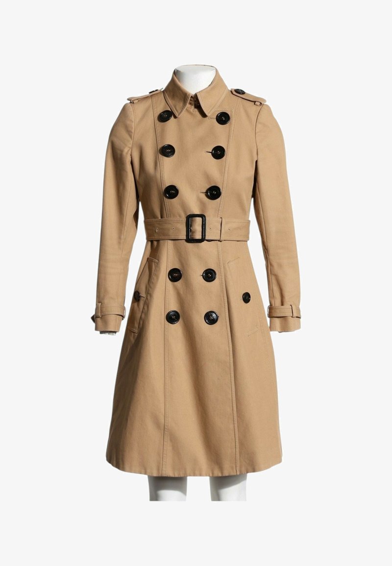 Trench-coat beige croisé avec boutons noirs, taille ceinturée, épaulettes aux épaules et brides aux poignets sur un mannequin sans tête.