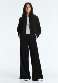 Femme debout les mains dans les poches, portant une veste noire à fermeture éclair, un pantalon large noir, un haut gris et des chaussures blanches sur un fond uni.